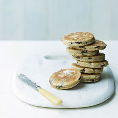 A stack of toasted cakes with raisins sits on a white marble board. A small butter knife with a light yellow handle rests beside the stack. The background is a soft white, creating a minimalist and airy setting.