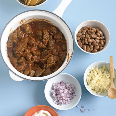 A bowl of beef chilli in a white saucepan with little bowls of grated cheese, chopped onion, beans and cream surrounding it