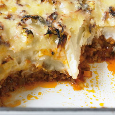 A dish of shepherds pie with a portion removed showing minced lamb and vegetables and a topping of mashed potato, leeks and cheese