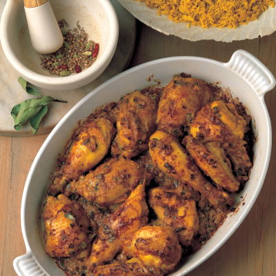 A white oval dish with pieces of spiced cooked chicken, a pestle and mortar next to it with ground spices and a plate of rice
