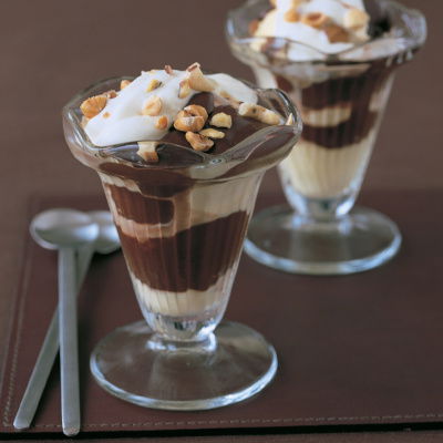Two glass sundae dishes layered with cream and chocolate with nuts sprinkled on top and two spoons on the side