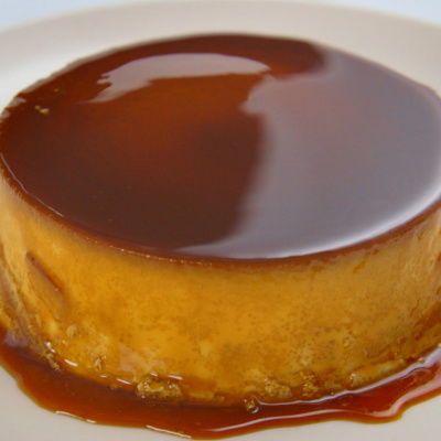 A smooth, glossy caramel flan sits on a white plate, its rich caramel sauce dripping down the sides and pooling around the base. The dessert has a golden-brown top and a creamy, custard-like texture.