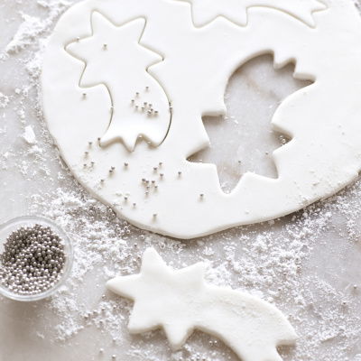 a picture of a circular sheet of white fondant icing on a white surface. three shooting stars have been cut out, a little dish of silver balls alongside and icing sugar on the top