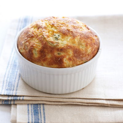 A golden-brown puff pastry with leaf designs on top, covering a white ceramic dish. It sits on a folded gray cloth atop a wooden board, with a silver spoon beside it.