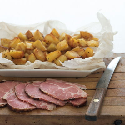 A picture of diced sauteed potatoes in a white dish sitting on a wooden board with sliced ham and a knife on the side