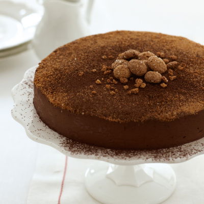 A chocolate torte on a white cake stand on a white napkins with red edging and a white jug with plates in the background