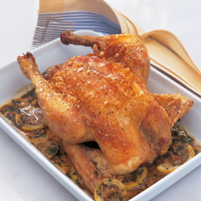 A cooked roast chicken in a white oven dish surrounded by the juices and slices of lemon