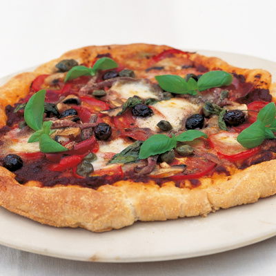 A cooked pizza on a white plate with a topping of basil, tomato, cheese and olives