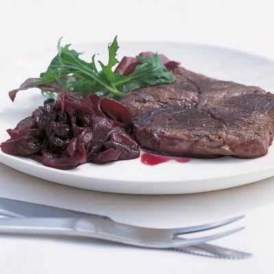 A grilled venison steak on a white plate with confit over the top and a garnish