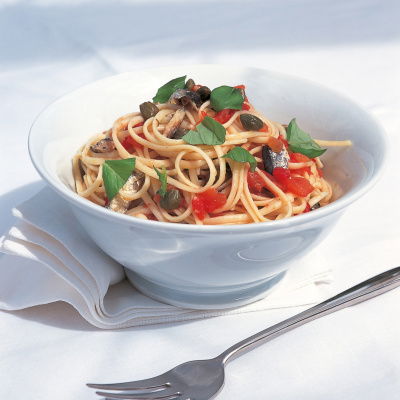 A white bowl of linguine pasta with tomatoes, capers, sardines and some torn basil to garnish. A fork is next to the bowl