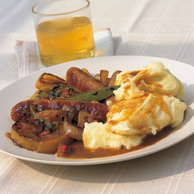 A picture of braised sausages with a pile of fluffy mash on a white plate with a glass of cider in the background
