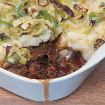 A white dish of shepherds pie with a portion removed showing minced lamb and vegetables and a topping of mashed potato, leeks and cheese