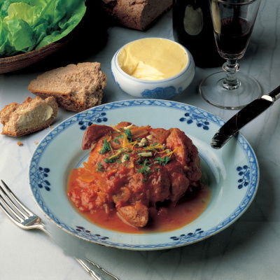 A picture of a blue and white china plate containing coked ossobuco with a knife and fork a glass of red wine and some bread and butter on the side