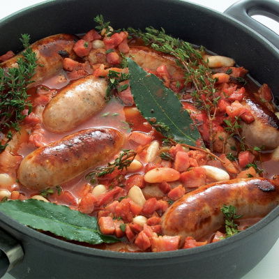 A picture of cooked cassoulet in a black casserole showing sausages, beans, bay leaves and tomatoes