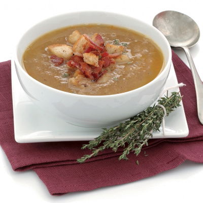 A picture of a white bowl on a folded napkin containg chestnut soup with croutons and bacon on the top and a little sprig of thyme next to it