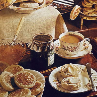 a table set with a china cup of tea and a saucer, a side plate with two muffins covered in butter, a plate of muffins, a jar of jam, a dish of marmalade and a roaring fire in the background