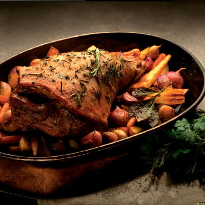 a cooked leg of lamb in a casserole dish with carrots and rosemary