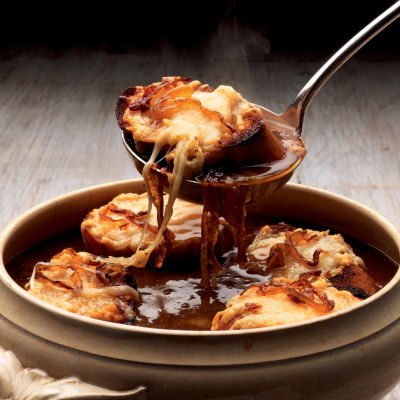 A picture of a soup bowl full of french onion soup with croutons covered in melted cheese and a ladle lifting out a whole crouton